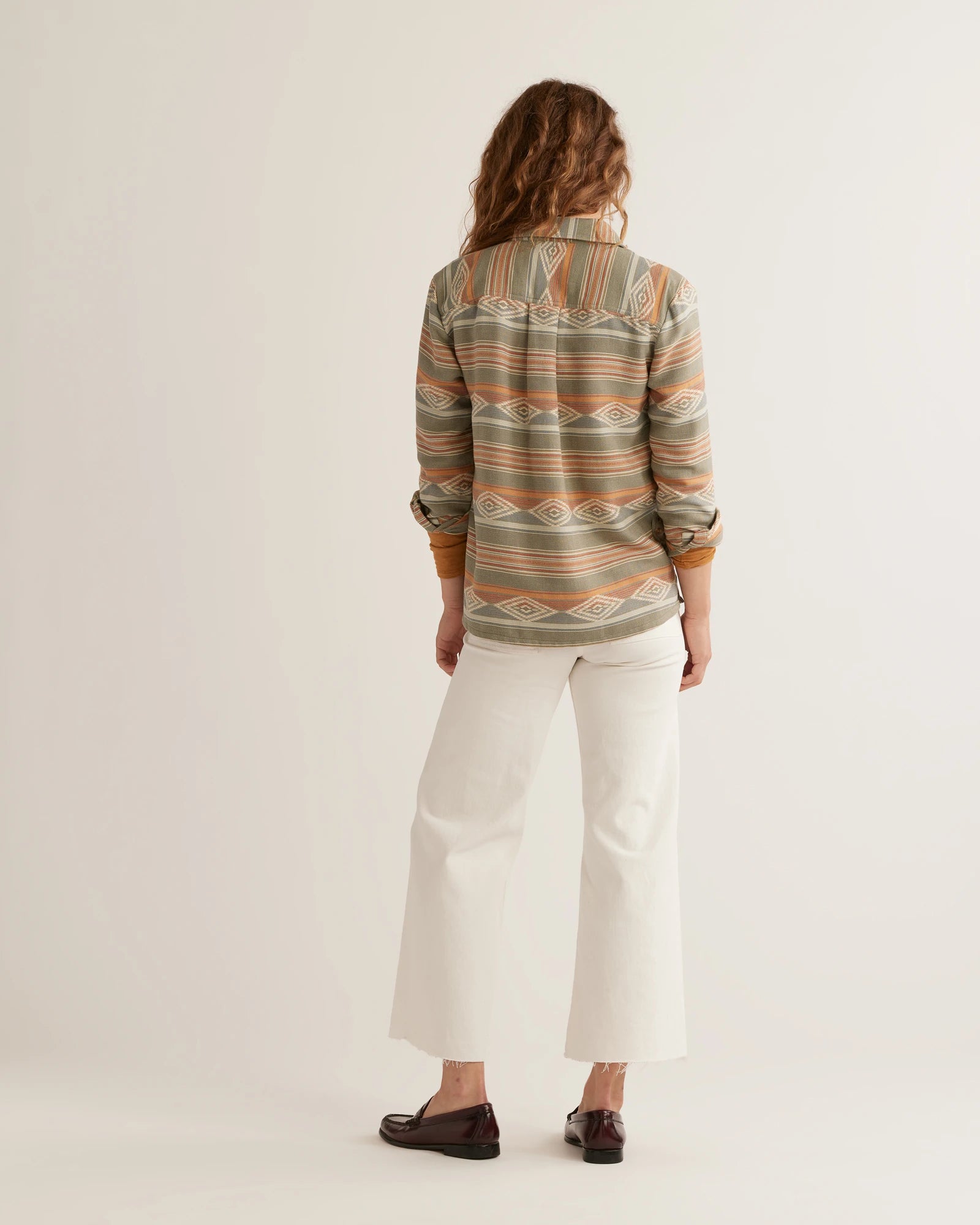 Back View of a woman wearing a Pendleton woven shirt with a geometric pattern in tones of beige, green, and brown, paired with white pants.