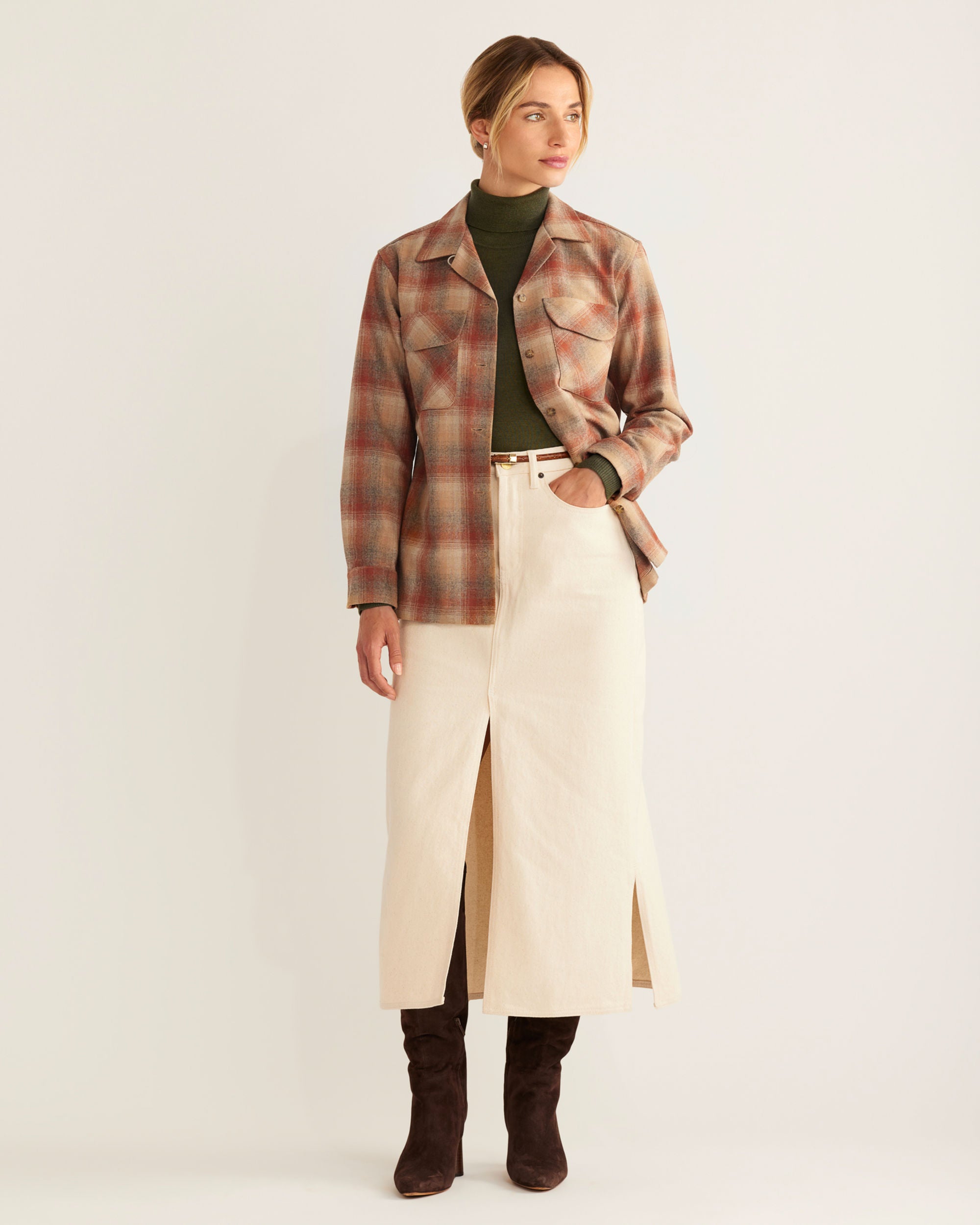 Woman wearing a plaid coat, green sweater, beige skirt, and brown boots on a white background
