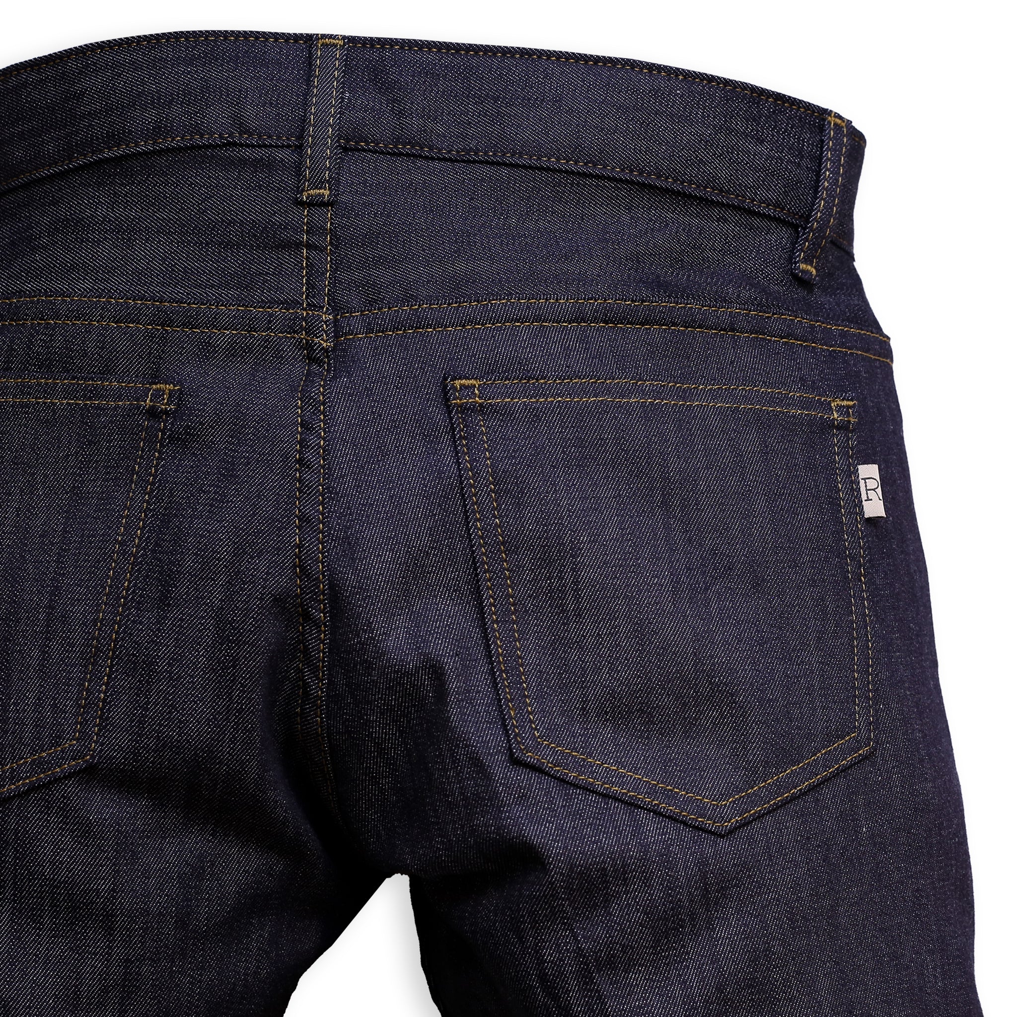 Back Pocket Detail of a pair of slim-fit blue denim jeans with a minimalist design, featuring a small 'R' logo on the back pocket.