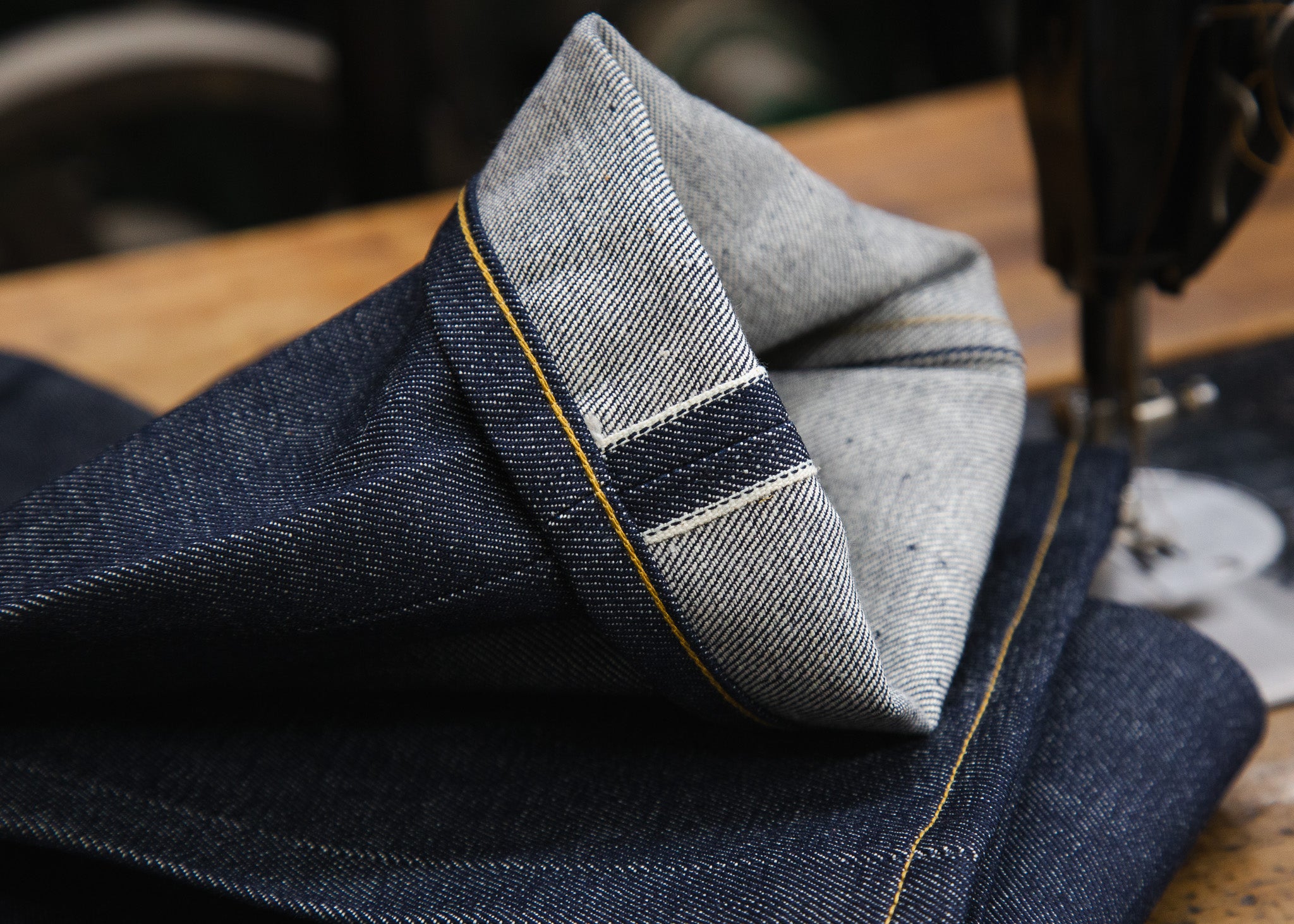 A close-up of Railcar selvedge denim folded on a sewing table beside an industrial machine, showing the rolled cuff and selvedge line